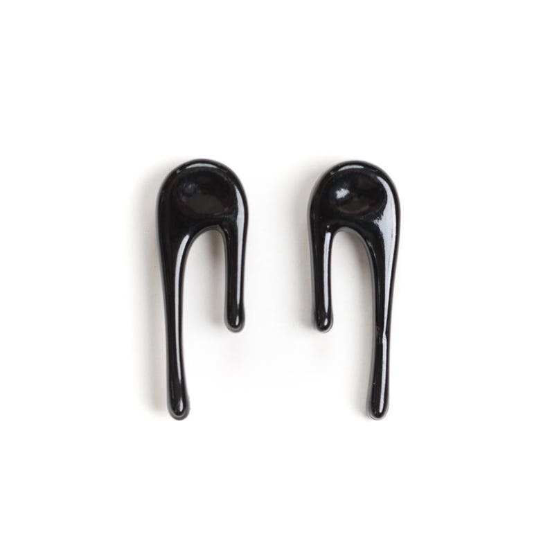 Puede incluir: Un par de pendientes negros abstractos en forma de gota. Los pendientes est&aacute;n hechos de un material liso y brillante.