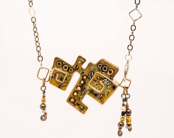 May include: A gold and silver necklace with a geometric pendant made of mixed materials. The pendant is a complex design with a brown, green, and gold color scheme. The necklace has a silver chain and a silver clasp. The pendant has a textured surface and is decorated with small beads and other embellishments.