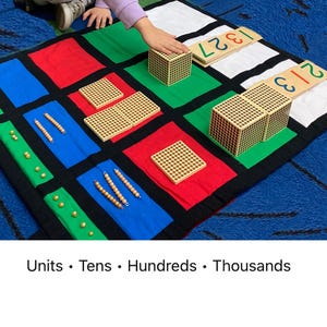 May include: A colorful educational mat with blocks and beads for learning math concepts. The mat is divided into squares of red, green, blue, and white. Wooden blocks and number cards are placed on the mat. The text "Units • Tens • Hundreds • Thousands" is at the bottom.