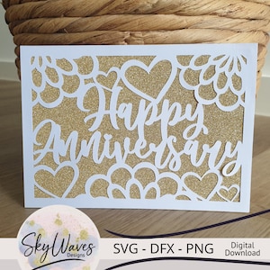 Op de afbeelding: Witte kaart met een gouden glitterachtergrond en een wit uitgesneden ontwerp van harten en bloemen. De tekst "Happy Anniversary" is geschreven in een wit cursief schrift.