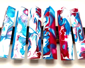 May include: Seven rectangular resin pieces with a marbled effect in white, blue, and red. The pieces are arranged on a dark wooden base. The colors swirl together, creating a unique pattern on each piece. The overall effect is vibrant and eye-catching.