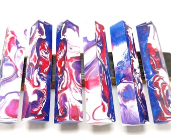 May include: Six rectangular resin blocks with a marbled design in red, white, and blue. The blocks are arranged side by side, showcasing a vibrant, swirled pattern. The blocks are on a dark wooden base.