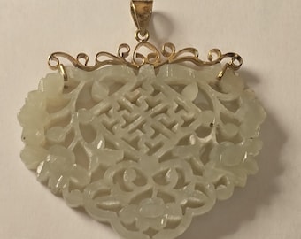 May include: A pale green jade pendant with a gold-colored bail and decorative filigree. The pendant features carved floral and geometric patterns. The jade is a light, translucent color. The pendant is likely for a necklace.