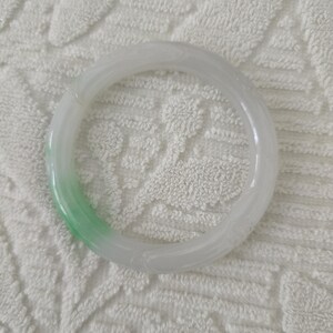 May include: A translucent jade bangle bracelet. The bracelet is primarily white with a gradient of light green. The smooth, circular design is a classic jewelry piece. The jade is polished and has a subtle sheen.