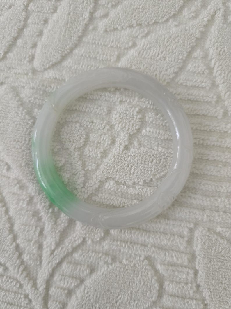 May include: A translucent jade bangle bracelet. The bracelet is primarily white with a gradient of light green. The smooth, circular design is a classic jewelry piece. The jade is polished and has a subtle sheen.