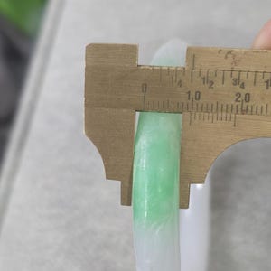 May include: A jade bangle with a gradient of white to light green. A brass caliper measures the width of the bangle. The bangle's width is approximately 0.4 inches.