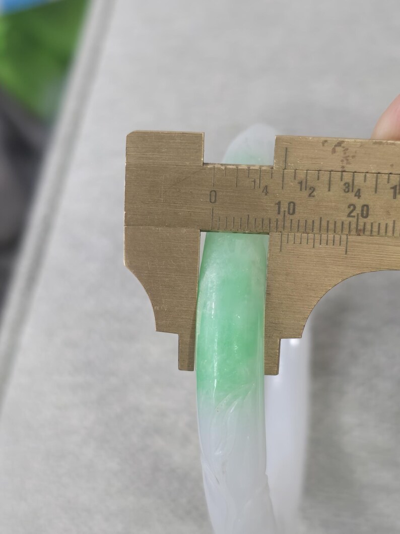 May include: A jade bangle with a gradient of white to light green. A brass caliper measures the width of the bangle. The bangle's width is approximately 0.4 inches.