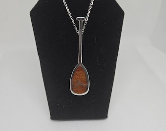 May include: A silver chain necklace with a wooden paddle pendant. The paddle is brown with a wave design carved into it.