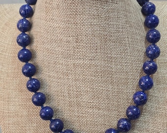 May include: A necklace made of dark blue lapis lazuli beads. The beads are round and have a speckled texture. The necklace is displayed on a brown fabric mannequin.