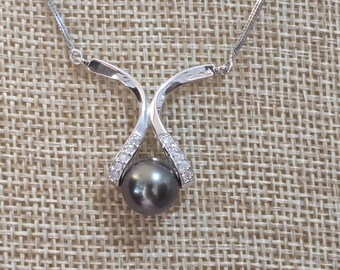 May include: A silver necklace featuring a dark gray pearl pendant. The pendant has a curved design with small, clear stones. The necklace chain is delicate and silver. The jewelry is displayed on a textured, tan surface.