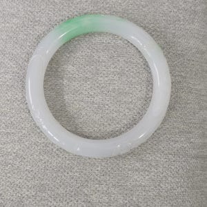 May include: A white jade bangle bracelet with a gradient of light green at the top. The bracelet has a smooth, polished surface and is carved with subtle floral patterns. The circular design is a classic jewelry style.