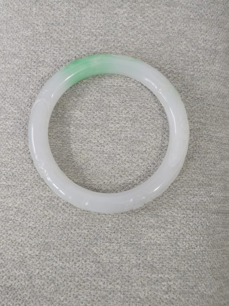 May include: A white jade bangle bracelet with a gradient of light green at the top. The bracelet has a smooth, polished surface and is carved with subtle floral patterns. The circular design is a classic jewelry style.