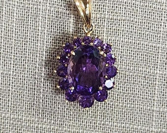 May include: A gold pendant featuring a large, oval-cut purple gemstone surrounded by smaller, round purple gemstones. The pendant has a gold bail for attaching to a chain. The gemstones are set in a gold setting.