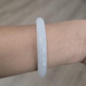 May include: A white jade bangle bracelet with carved floral details. The bracelet is a light, translucent color and is worn on a wrist. The jade is smooth and polished, reflecting light.