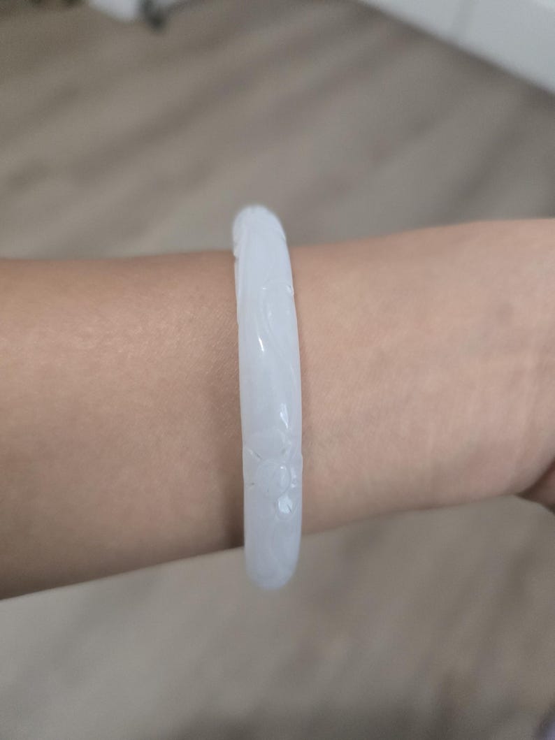 May include: A white jade bangle bracelet with carved floral details. The bracelet is a light, translucent color and is worn on a wrist. The jade is smooth and polished, reflecting light.