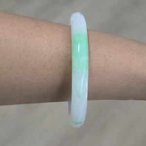 May include: A jade bangle bracelet with a smooth, polished surface. The bracelet features a gradient of colors, transitioning from white to a vibrant green. Carved floral details add texture to the white sections.