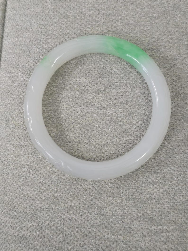 May include: A jade bangle bracelet, featuring a smooth, polished surface. The bracelet transitions from a white hue to a vibrant green. This piece of jewelry is a circular, solid band, showcasing a natural stone aesthetic.