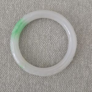 May include: A white jade bangle with a touch of green. The smooth, circular bracelet features subtle carved details. The jade is a pale white with a gradient of green on one side, creating a striking contrast.
