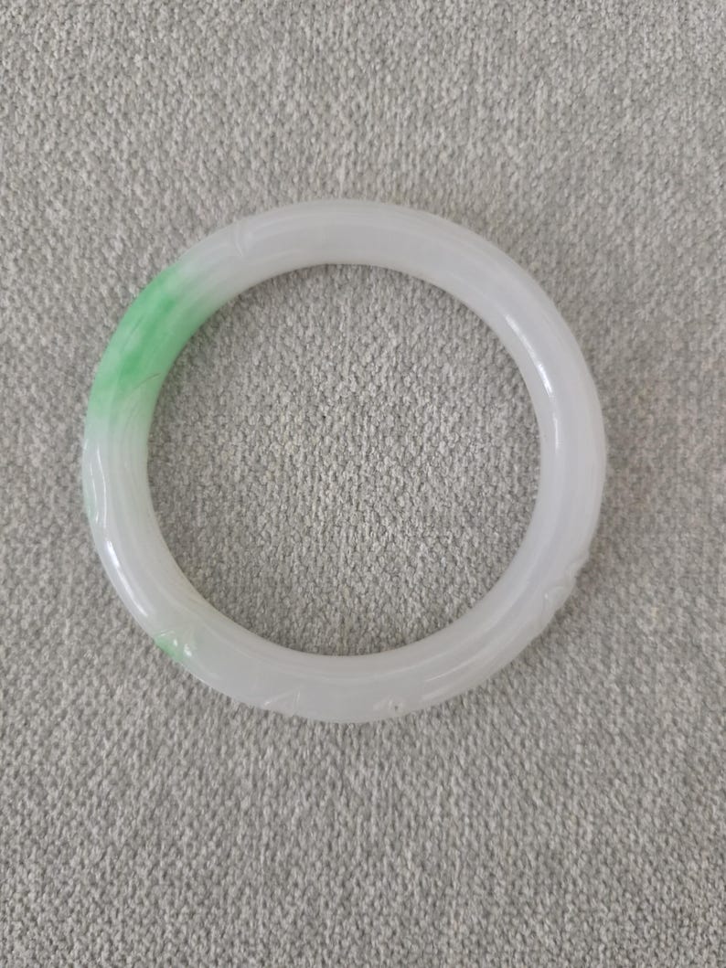 May include: A white jade bangle with a touch of green. The smooth, circular bracelet features subtle carved details. The jade is a pale white with a gradient of green on one side, creating a striking contrast.