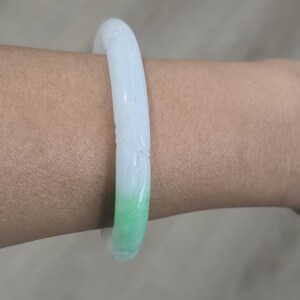 May include: A jade bangle bracelet with a carved floral design. The bracelet transitions from white to a vibrant green hue. The smooth, polished surface reflects light, highlighting the intricate details of the carving.