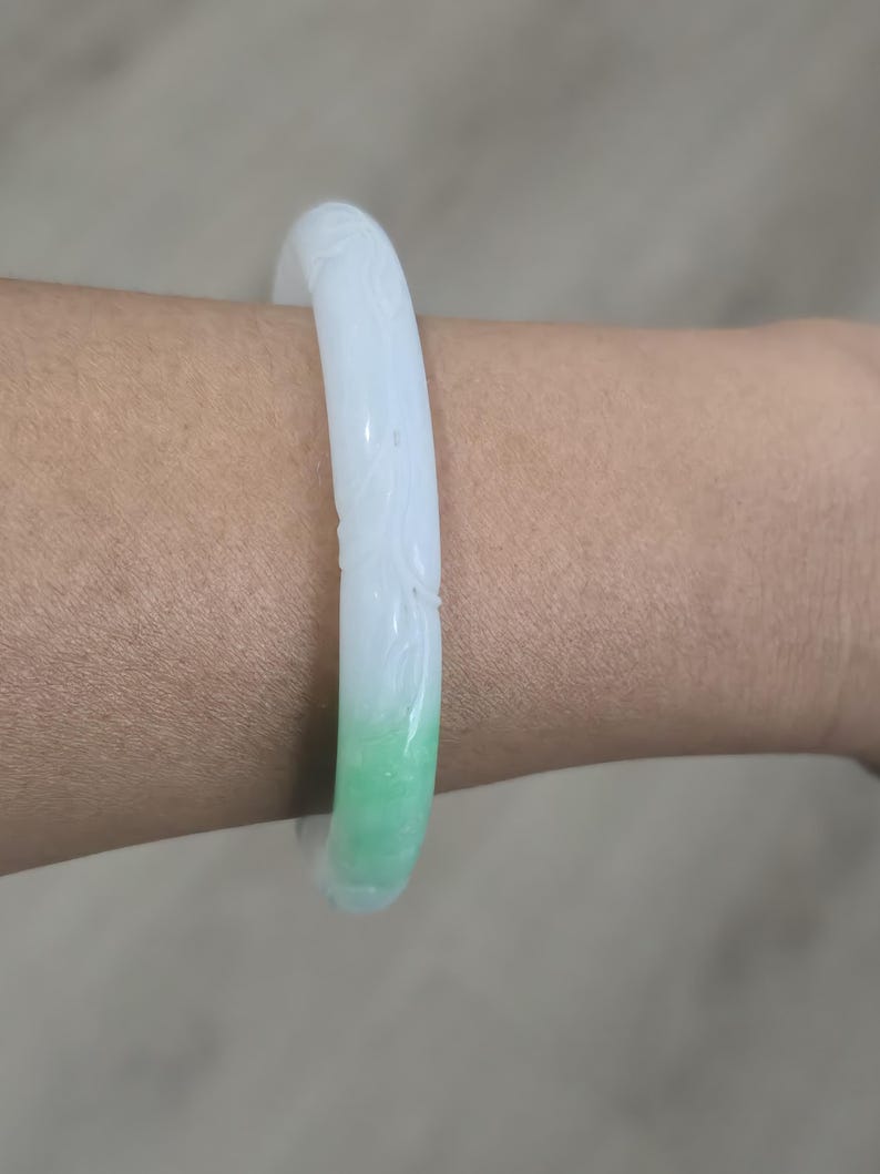 May include: A jade bangle bracelet with a carved floral design. The bracelet transitions from white to a vibrant green hue. The smooth, polished surface reflects light, highlighting the intricate details of the carving.