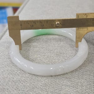 May include: A white jade bangle with carved floral details. A brass caliper measures the bangle's diameter. The jade has a slight green hue in one section. The bangle is approximately 3 inches in diameter.