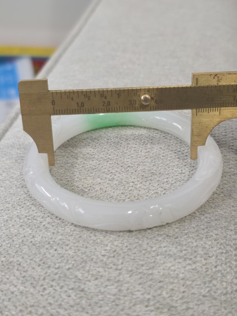 May include: A white jade bangle with carved floral details. A brass caliper measures the bangle's diameter. The jade has a slight green hue in one section. The bangle is approximately 3 inches in diameter.