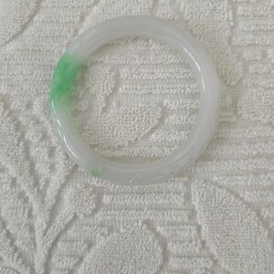 May include: A translucent jade bangle bracelet. The bracelet is primarily white with a section of light green. The smooth, circular design is a classic jewelry piece. The bracelet is shown on a white, textured surface.