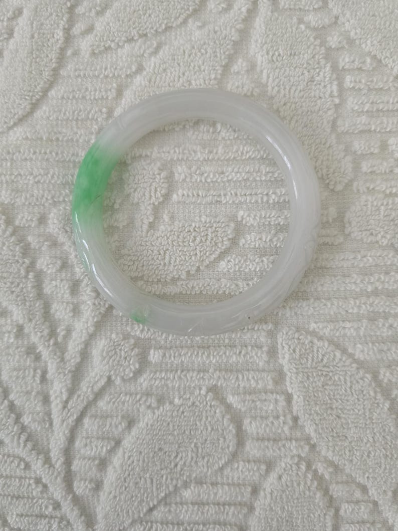 May include: A translucent jade bangle bracelet. The bracelet is primarily white with a section of light green. The smooth, circular design is a classic jewelry piece. The bracelet is shown on a white, textured surface.