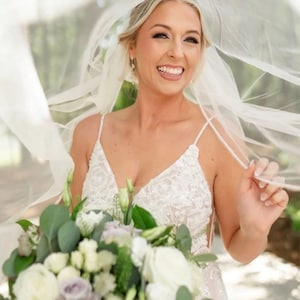 May include: A bride smiles while holding a bouquet of white and purple flowers, wearing a white lace wedding dress and a white veil.