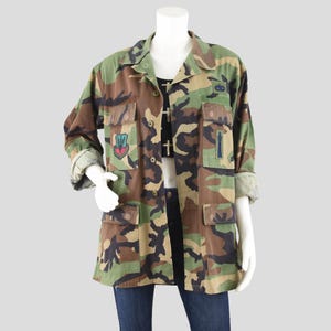 May include: Camouflage print jacket with multiple pockets and patches. The jacket features a button-up front, rolled-up sleeves, and a collared neckline. The color palette includes shades of green, brown, and black.