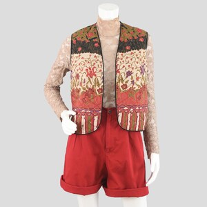 May include: A sleeveless vest with a floral pattern in red, green, and cream colors. The vest has a black border and is paired with red shorts. The outfit is displayed on a mannequin.