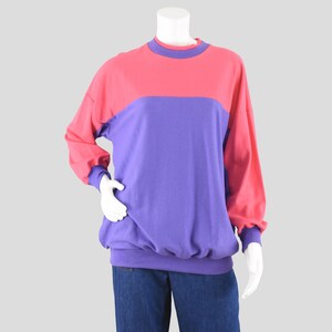 May include: A color block sweatshirt in shades of pink and purple. The top section is pink, and the lower section is purple. The sleeves and cuffs are pink with purple trim. The sweatshirt is displayed on a mannequin.