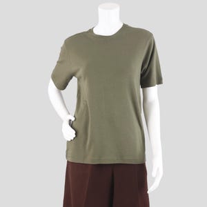 May include: Olive green crew neck t-shirt with short sleeves. The shirt is made of a soft, solid-colored fabric. The shirt is displayed on a mannequin with brown pants.