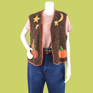 May include: Brown textured vest with orange trim, featuring appliqued stars, a crescent moon, and pumpkins. The vest is worn over a pink t-shirt and blue jeans, with a brown belt. The overall theme is autumn and fall.