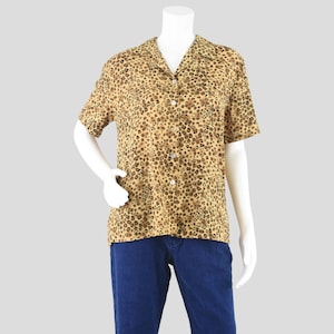 May include: A short-sleeved button-down shirt with a leopard print pattern in shades of tan and brown. The shirt has a classic collar and is paired with blue denim jeans. The shirt is displayed on a mannequin.