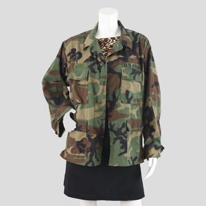 May include: Camouflage pattern jacket with button-up front, two chest pockets, and the text "U.S. ARMY" on the right chest. The jacket is displayed on a mannequin, paired with a black skirt and a leopard print top.