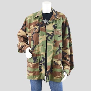 May include: A camouflage pattern jacket with multiple pockets and button closures. The jacket features a mix of green, brown, and black hues. The jacket is worn over a black t-shirt and blue jeans.