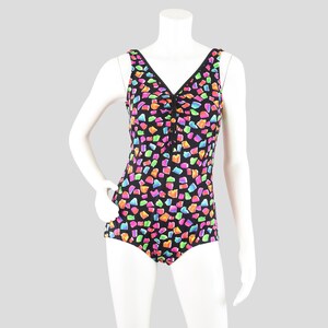 May include: A black one-piece swimsuit with a V-neck and a button closure. The swimsuit is covered in a colorful, abstract pattern of pink, orange, green, and blue shapes. The swimsuit is displayed on a white mannequin.