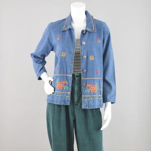 May include: A light blue denim jacket with floral embroidery in red, orange, and green. The jacket has a collared neckline, button-up front, and three-quarter sleeves. The jacket is displayed on a mannequin with green corduroy pants and a striped shirt.