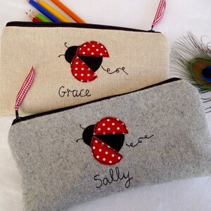 May include: Two personalized pencil cases with ladybug designs. The top case is beige with the name "Grace" embroidered on it. The bottom case is gray with the name "Sally" embroidered on it.