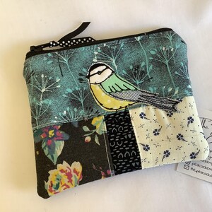 May include: A rectangular fabric zipper pouch featuring a patchwork design. The top section is teal with a white botanical print and a bird appliqué. The bottom section consists of three panels: a black floral print, a black geometric print, and a cream floral print.