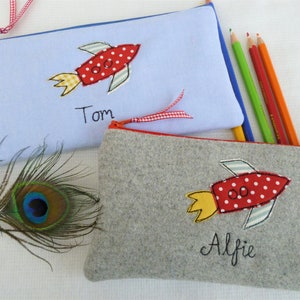 Peut inclure: Deux trousses à crayons personnalisées avec un motif de fusée. La trousse du haut est bleu clair avec le nom "Tom" brodé dessus. La trousse du bas est grise avec le nom "Alfie" brodé dessus.