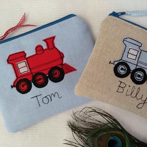 May include: Two zippered pouches with embroidered train designs. One pouch is light blue with a red train and the name "Tom". The other is beige with a blue train and the name "Billy". Both have blue zippers.