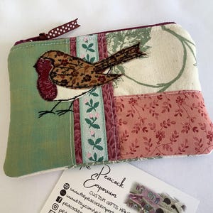 May include: A rectangular fabric zipper pouch featuring a patchwork design with green, cream, and pink floral patterns. An appliqued brown and red bird is stitched onto the front. A brown ribbon with white polka dots is attached to the zipper pull.