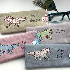 May include: Four personalized glasses cases with embroidered horses and names. The cases are made of different fabrics, including linen, pink, and gray. The names on the cases are Ada's Glasses, Mum, Eve's Sun, and Holly's Specs.
