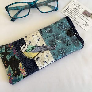 Puede incluir: Un estuche de tela para gafas con diseño de patchwork, una aplicación de pájaro azul, motivos florales y cierre de botón negro. Junto al estuche hay unas gafas de lectura de color azul cerceta y una tarjeta de The Peacock Emporium.