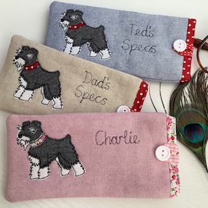 Könnte beinhalten: Drei personalisierte Brillenetuis mit gestickten Hunden und Text. Die Etuis sind aus einem weichen, strukturierten Stoff gefertigt. Das erste Etui ist beige mit roten Punkten und trägt die Aufschrift "Dad's Specs". Das zweite Etui ist blau mit roten Punkten und trägt die Aufschrift "Ted's Specs". Das dritte Etui ist rosa mit floralem Besatz und trägt die Aufschrift "Charlie".