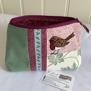 May include: A fabric zip pouch featuring a patchwork design with a green panel, a dark pink floral panel, a light blue ribbon with pink flowers, and a cream panel with a brown bird appliqué. The interior is lined with dark pink fabric.