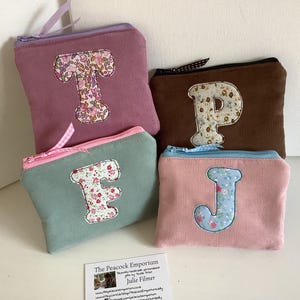 May include: Four corduroy zipper pouches in shades of pink, brown, and teal. Each pouch features a floral-patterned letter applique: T, P, F, and J. The pouches have matching coloured zippers and ribbon pulls.